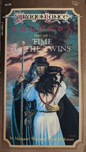 The cover of Time of the Twins. A black-robed, bronze-skinned mage stares directly at the viewer. In one hand he holds a magic staff; the other arm is around the waist of a black-haired young woman in white robes. She seems distracted, staring off to the side with a concerned expression. Behind them is a sky full of roiling clouds.